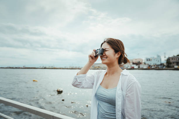A side shot of smiling Asian woman using a one-time-use camera to take city photo outdoors next to the sea