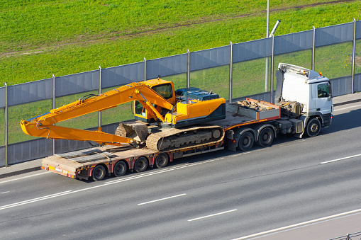 Heavy new yellow excavator long boom bucket on transportation truck with long trailer platform on the highway in the city