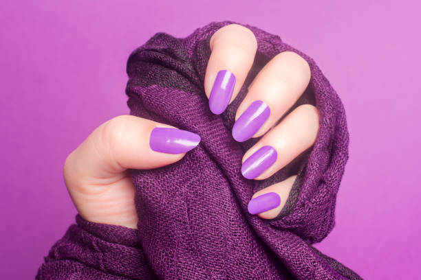 Female hand with purple or pink nails is holding purple textile on pink background, manicure and nail care concept.