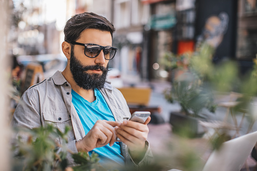 Portrait of a long beard middle eastern ethnicity hipster young man texting with smartphone and working laptop at outdoor urban coffee shop