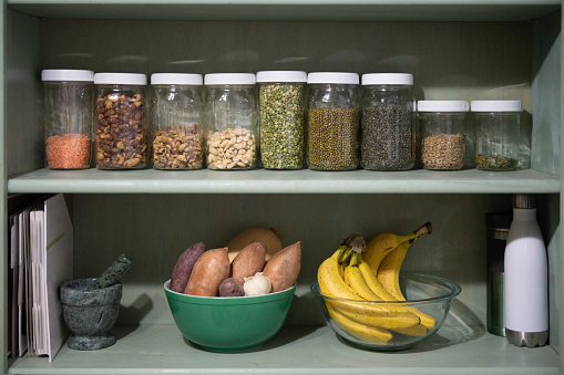 This is a candid photograph of a kitchen pantry shelf lined with jars full of vegan dry food including quinoa, lentils, green peas, sweet potatoes, bananas, and beans at home.