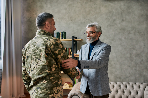 Mature psychologist smiling, shaking hands with middle aged military man after therapy session. Soldier suffering from depression, psychological trauma. PTSD concept. Focus on aged man