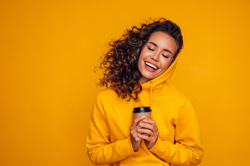 Happy young cheerful girl laughing and holding cup of coffee on colored yellow background