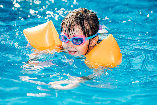Little black-haired girl smiling and swimming in the pool wearing sleeves and swimming goggles