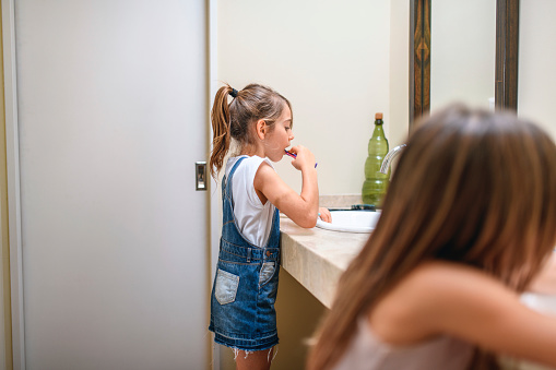 Young girl with hair in ponytail standing next to her sister at family bathroom sink and brushing her teeth.