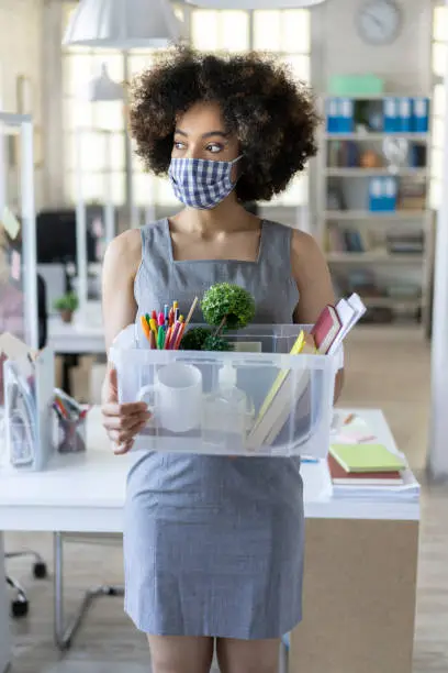 Unhappy afro business woman with surgical medical mask going out with box container, looking at camera and feeling looser.