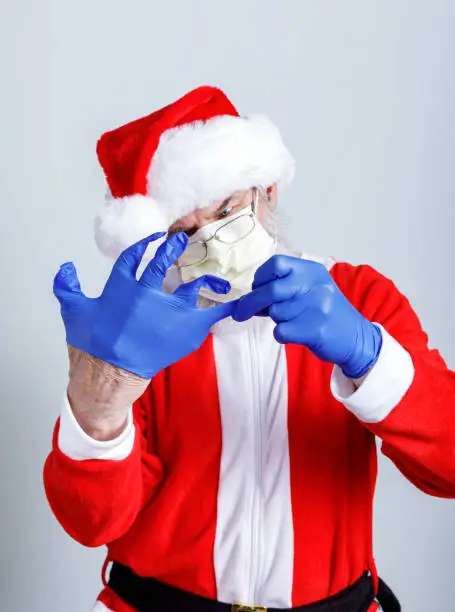 Santa Claus wearing a Covid-19 coronavirus pandemic PPE (Personal Protective Equipment) face mask is fumbling with his blue nylon gloves - trying to get his fingers into the proper places.