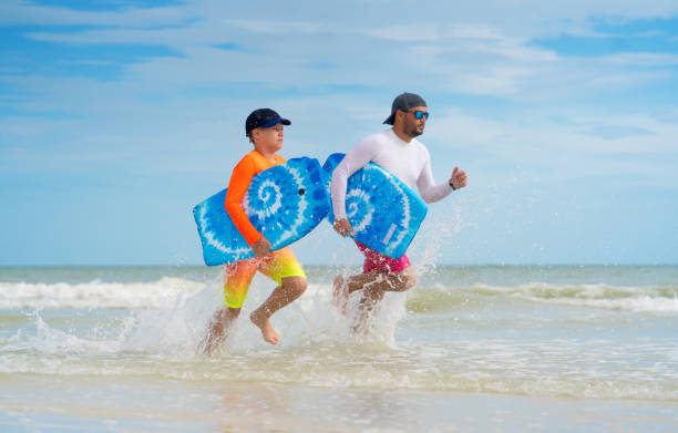 istock Father and son running with skimboards on shallow waters 1262117436