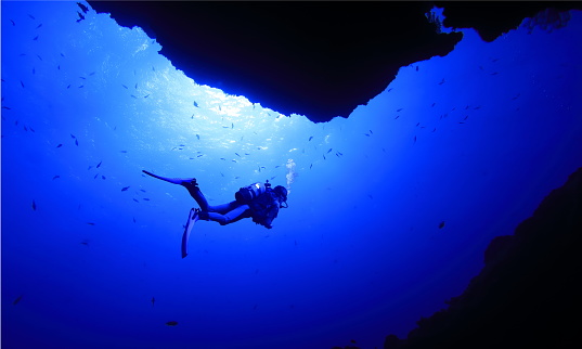 A diver swimming at the exit of the Queen's room in a seabed cave at a diving spot in Miyakojima City, and sunlight pouring overhead.