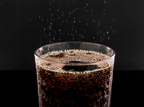 Ice Cola with splashes and bubbles. Drink in a transparent glass on a black background. Refreshing and thirst-quenching concept.