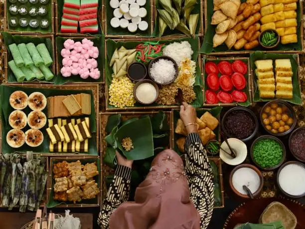 Top view of a market stall selling traditional Indonesian sweet and savory snacks. The snacks are assortment of traditional Javanese, Sundanese, Manadonese, Peranakan, Chinese-influenced and also Western-influenced. Here seen the lady seller is scooping snack called Tiwul (Javanese dried cassava granules) from a tray; her other hand is holding a traditional banana leaf containter called pincuk. The lady seller is wearing Javanese batik clothing with a hijab.