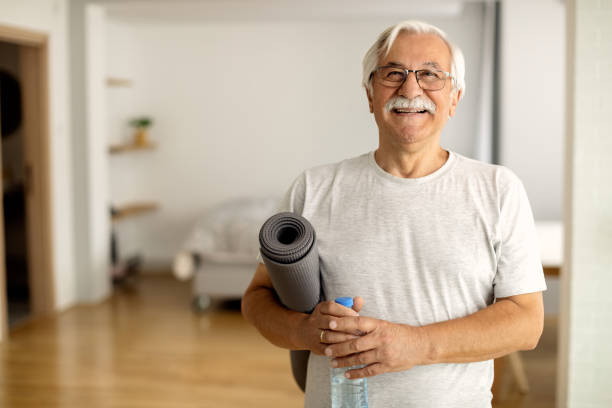 Exercising is the key of his vitality! Happy senior man holding bottle of water and exercise mat in the living room and looking at camera. home workout stock pictures, royalty-free photos & images