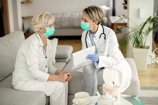 La doctora y su paciente senior usan máscaras protectoras para la cara durante una visita a casa. photo