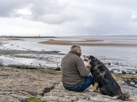 Man and dog sitting on rocky shore rear view