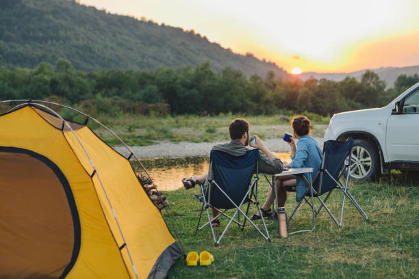 pareja sentada en sillas de campamento mirando al atardecer sobre el río en las montañas - camping fotografías e imágenes de stock