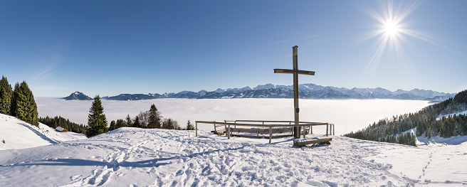 Summit Cross on snow mountain with blue Sky and Sun. Amazing view to Mountain Range above inversion foggy cloud layer. Ofterschwanger Horn, Alps, Allgäu, Bavaria, Germany.