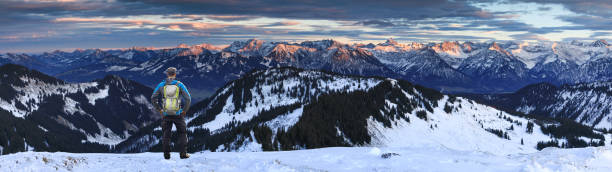 山岳地帯と劇的な空の素晴らしい夕日アルペングローパノラマを眺めながら、雪の冬の山に立ってバックパックを持つハイカーマン。リードベルガーホルンからアルゴイアルプスまでの眺め� - copy space alpenglow winter mountain range ストックフォトと画像