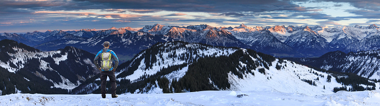 Hiking Man with Backpack standing in snow on winter mountain with view to amazing sunset alpenglow panorama of snow covered mountain ranges and dramatic clouds in cold winter. View from Riedbergerhorn to Allgäu Alps Mountains. Bavaria, Germany.