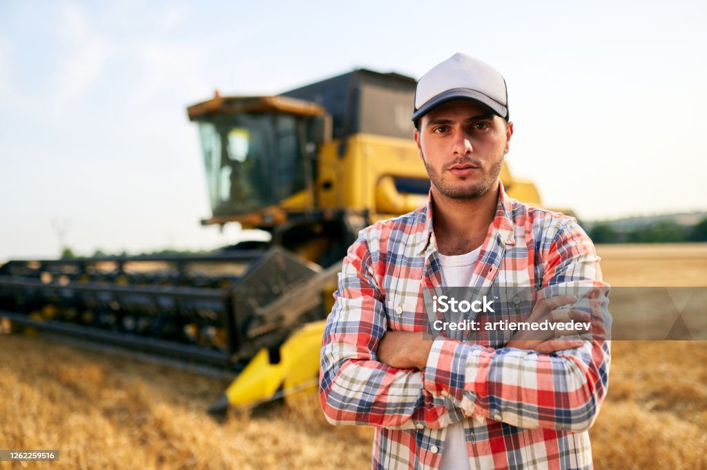 Portrait of proud harvester machine driver with hands crossed on chest. Farmer standing at his combine. Agronomist looking at camera. Rancher at harvesting work on stubble of harvested wheat field Portrait of proud harvester machine driver with hands crossed on chest. Farmer standing at his combine. Agronomist looking at a camera. Rancher at harvesting work on stubble of harvested wheat field. Farmer Stock Photo