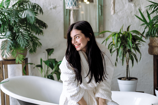 Happy woman spending morning at bathroom, looking aside, smiling wide and sitting on bath in bathrobe. Dreamy young adult female with healthy and brunette floating hair
