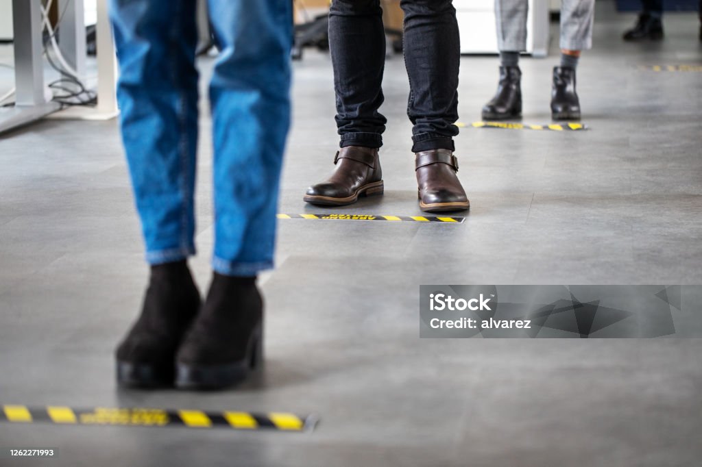 Business people standing behind social distancing signage on office floor Low angle shot of business people standing behind social distancing signage on office floor. People in queue at office for safe entry checks post corona virus pandemic. Coronavirus Stock Photo