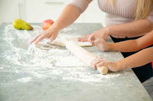 Hands of mom and daughter rolling dough on kitchen table. Girl and her mother baking bread or cake together. Closeup, cropped shot. Family cooking concept