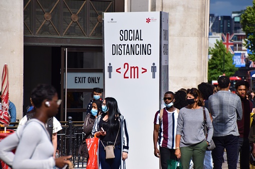 London, United Kingdom - July 28 2020: People wearing protective face masks walk past a social distancing sign on Oxford Circus