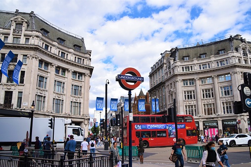 London, UK - May, 2023. People crossing street and sit on steps of monument at Piccadilly Circus Street in London during daytime in springtime with Piccadilly Circus Shopping center behind and huge advertisement board. British Flag and Coronation Flag  Landscape format.