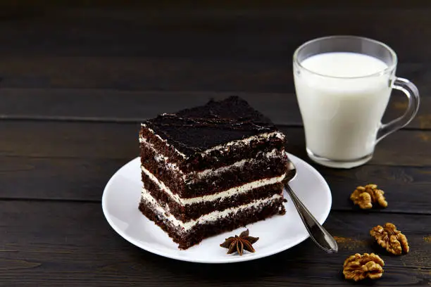 A piece of chocolate brownie cake with walnuts and a mug of milk on a dark wooden background.