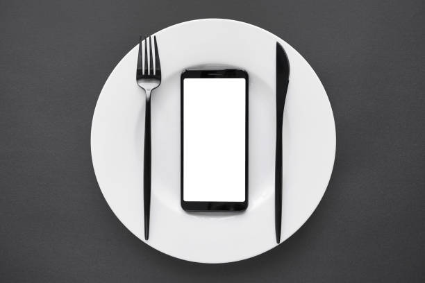 istock Smart phone mock up screen on white plate fork and knife black background table, food delivery app concept. Take away business restaurant meal online mobile application deliver order flat lay top view 1262278847