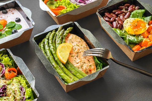 menú de catering de alimentos saludables en loncheras de pescado y paquetes de verduras como entrega diaria de mensajería plan de dieta de comidas con tenedor aislado sobre fondo de mesa negro. quitar el concepto de pedido de contenedores. - food healthy eating healthy lifestyle meal fotografías e imágenes de stock