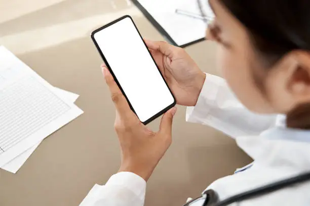 Photo of Indian female doctor holding in hands using cell phone app mock up white screen, over shoulder closeup view. Healthcare telemedicine online consultation, remote mobile medical smartphone application