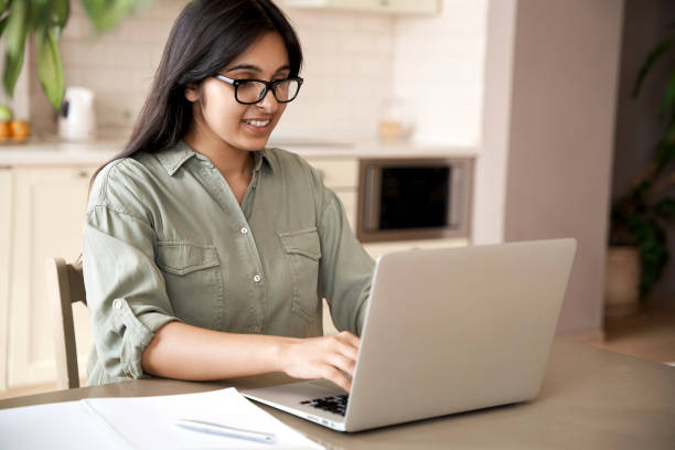 Smiling indian young adult woman wearing glasses typing on laptop computer working at home office sitting at table. Happy female professional freelancer student studying online using notebook pc. Smiling indian young adult woman wearing glasses typing on laptop computer working at home office sitting at table. Happy female professional freelancer student studying online using notebook pc. india train stock pictures, royalty-free photos & images