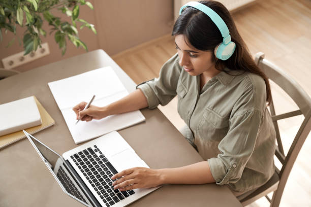 Indian girl student wear headphones learning online watching webinar class looking at laptop computer elearning remote lesson making notes or video calling virtual conference meeting teacher at home. Indian girl student wear headphones learning online watching webinar class looking at laptop computer elearning remote lesson making notes or video calling virtual conference meeting teacher at home. virtual college education stock pictures, royalty-free photos & images