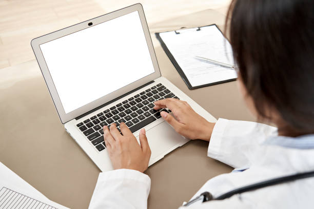 istock Indian female doctor wear white coat typing using laptop computer mock up white screen browsing internet sit at work desk. Healthcare medical e health website tech concept. Close up over shoulder view 1262283527