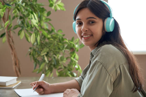sourire heureux jeune femme indienne de fille d’adolescence utilisant des écouteurs regardant l’appareil-photo étudiant à la maison écoute des notes d’écriture d’apprentissage à distance d’écoute fonctionnant à la table au bureau à la ma - 7652 photos et images de collection