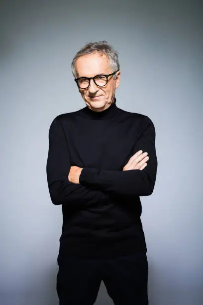 Fashion portrait of elegant senior man wearing black turtleneck and trousers, standing with arms crossed and looking at camera. Studio shot, grey background.