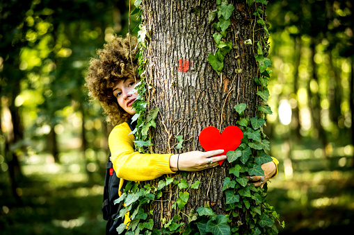 woman hand in the woods puts a heart shape on the trunk to tell us that every tree has a heart. Background of pines forest. Earth day concept. Together save the planet from deforestation. Girl sharing her heart with the tree.