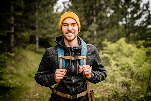 Young handsome man hiker with yellow cap and backpack walking through pine tree forest