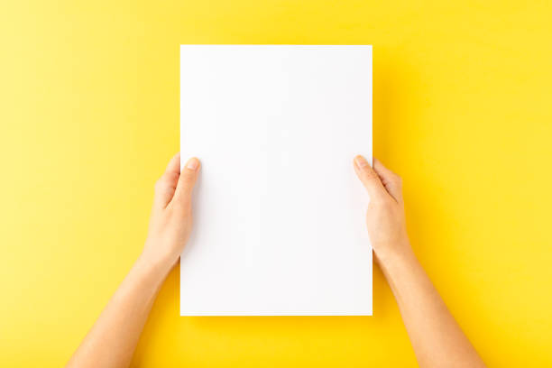 istock Young woman’s hands showing blank paper sheet on yellow desktop. Top view 1262294895