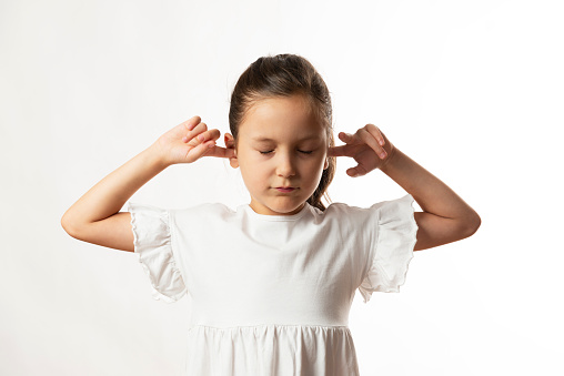 Little girl in white dress is having her fingers in ears. She does not want to hear anything.