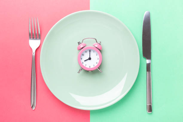 istock Pink alarm clock, fork, knife and empty plate on colored paper background. Intermittent fasting concept 1262299415