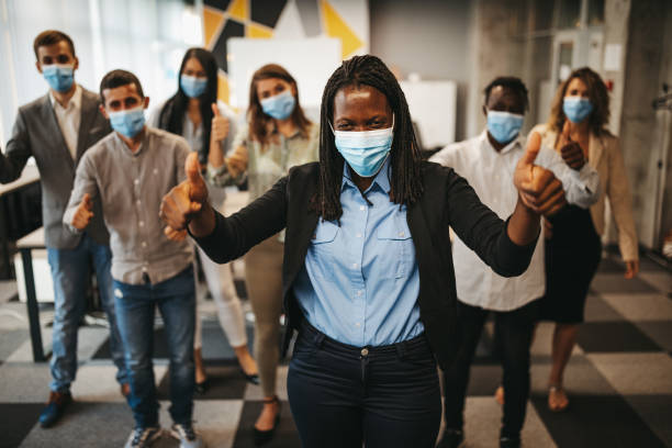 Group of business people with protective face masks celebrating success Cheerful multi-ethnic group of young business people with protective face masks standing and rejoice with thumbs up in modern office, celebrating successfully completed work thumbs up photos stock pictures, royalty-free photos & images