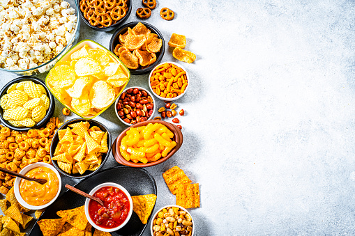 Party food: assortment of salty snacks in bowls shot from above on abstract gray table. The composition includes potato chips, popcorn, nacho chips and salsa, corn bugles, pretzels, peanut, cheese sticks and others. The composition is at the left of an horizontal frame leaving useful copy space for text and/or logo at the right. Predominant colors are yellow and gray. High resolution 42Mp studio digital capture taken with SONY A7rII and Zeiss Batis 40mm F2.0 CF lens