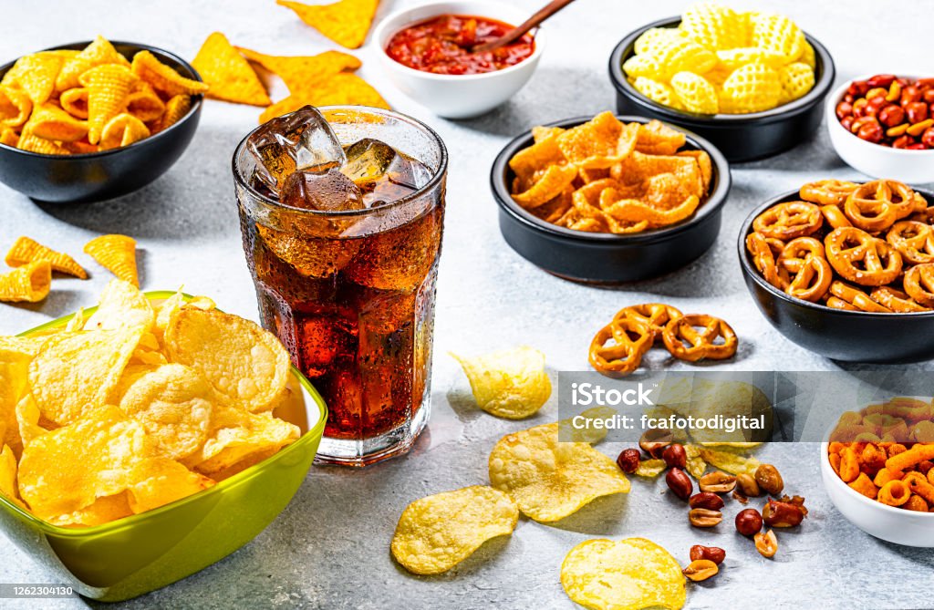 Cola soda glass and salty snacks shot on gray table Unhealthy food: cola soda glass and various salty snacks shot on gray table. The composition includes potato chips, pretzels, peanut, corn bugles, nacho chips and others. Predominant colors are yellow and gray. High resolution 42Mp studio digital capture taken with Sony A7rII and Sony FE 90mm f2.8 macro G OSS lens Snack Stock Photo