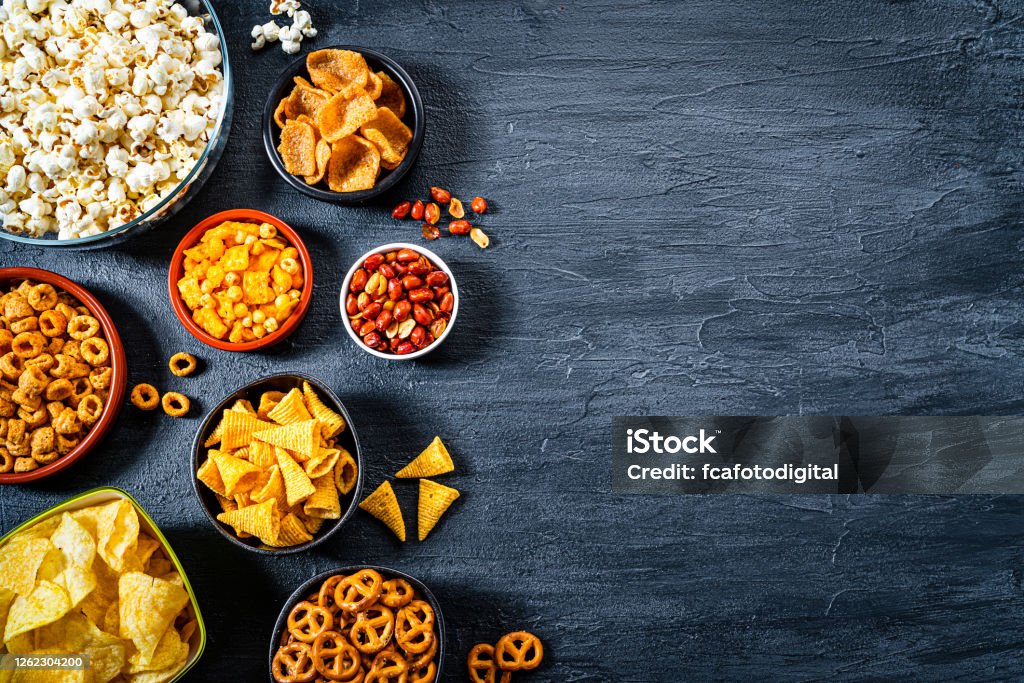 Salty snacks assortment shot from above on dark slate table. Copy space Party food: assortment of salty snacks in bowls shot from above on dark slate table. The composition includes potato chips, popcorn, corn bugles, pretzels, peanut, cheese sticks and others. The composition is at the left of an horizontal frame leaving useful copy space for text and/or logo at the right. Predominant colors are yellow and black. High resolution 42Mp studio digital capture taken with SONY A7rII and Zeiss Batis 40mm F2.0 CF lens Snack Stock Photo