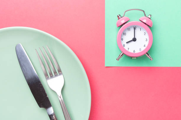 istock Pink alarm clock, fork, knife and empty plate on colored paper background. Intermittent fasting concept 1262306019