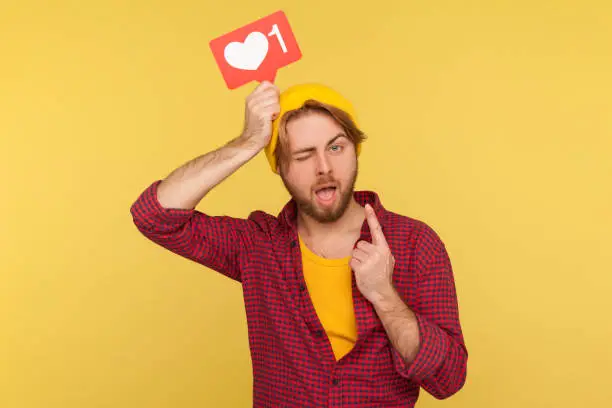Photo of Optimistic hipster guy in checkered shirt holding social network Heart Like icon over head and raising finger