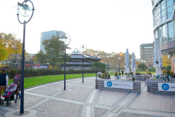 People, a restaurant and subway station Friedensplatz in Dortmund in autumn day. A woman and a man are walking with baby buggy in left area. In background is subway station. In right are are a few people at tables of a restaurant.