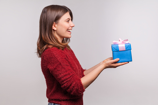 Please, take this christmas present! Side view, happy girl in shaggy sweater giving decorated gift box and greeting, congratulating on new year, x-mas holidays. studio shot isolated on gray background